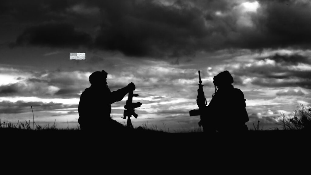 Two soldiers with their weapons sitting on a hill at dusk.